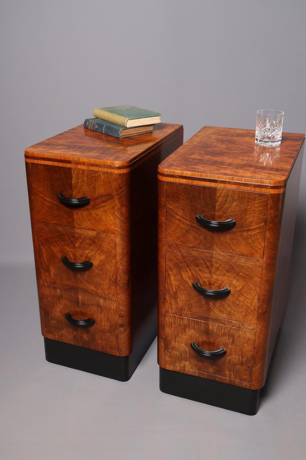 A matching pair of tall Art Deco burr walnut bedside cabinets with three drawers, dark curved handles, and ebonised plinth bases. View XI