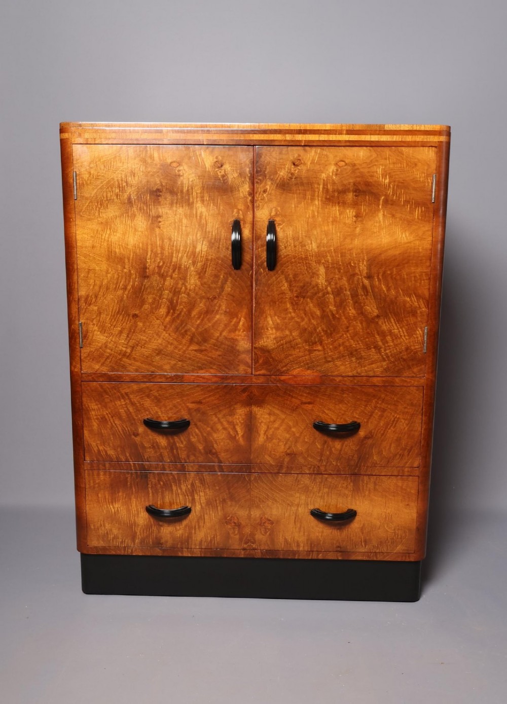 An Art Deco burr walnut compactum wardrobe featuring upper double doors, two long lower drawers, black curved handles, and an ebonised plinth.