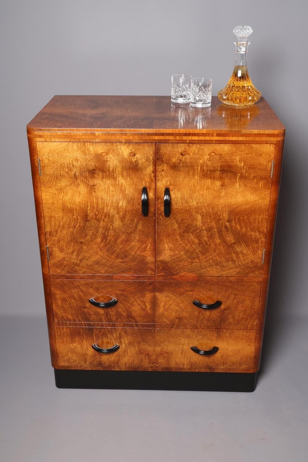 An Art Deco burr walnut compactum wardrobe featuring upper double doors, two long lower drawers, black curved handles, and an ebonised plinth. View II