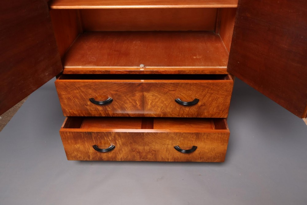 An Art Deco burr walnut compactum wardrobe featuring upper double doors, two long lower drawers, black curved handles, and an ebonised plinth. View X