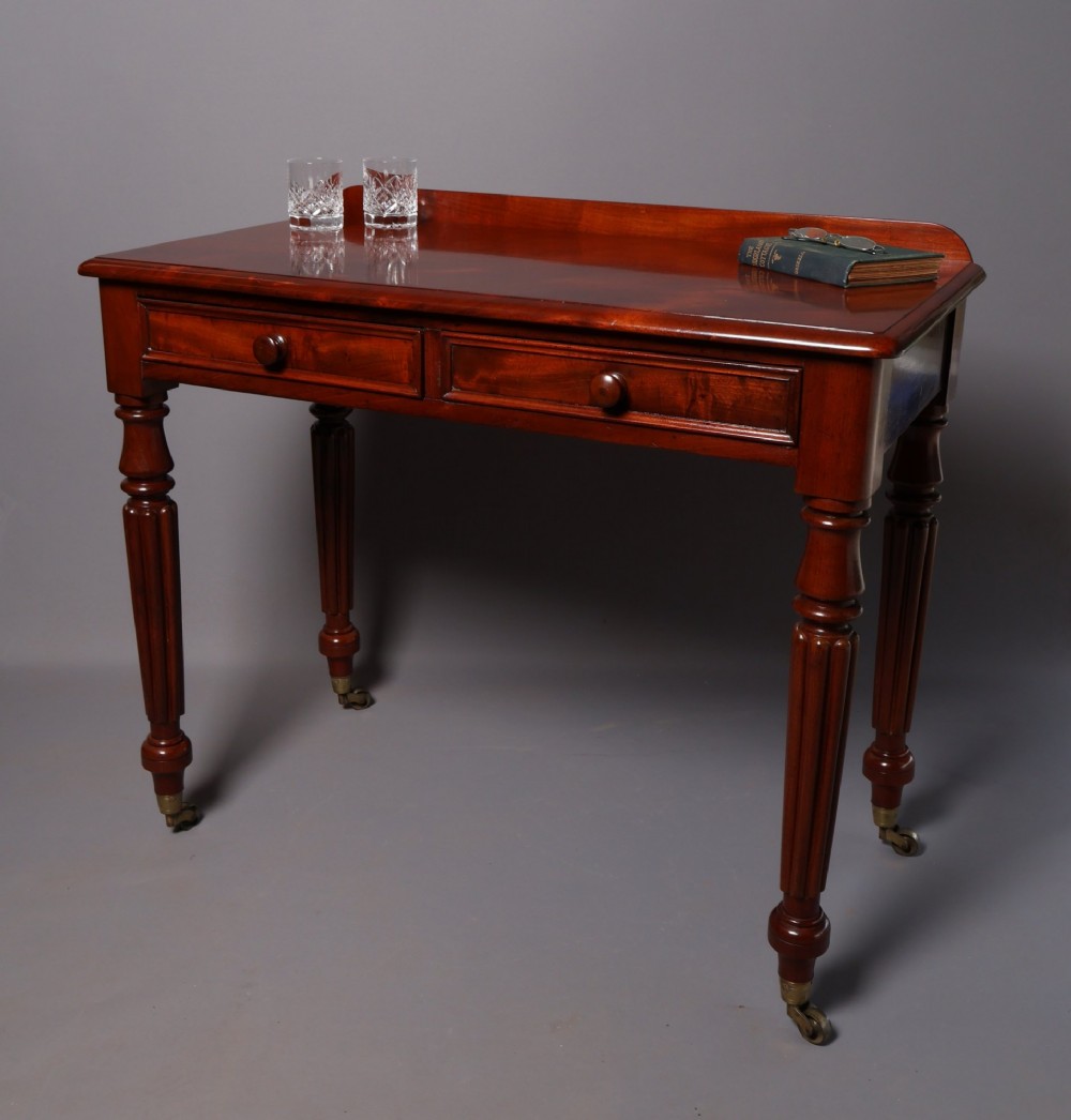 A Victorian mahogany writing table with a low gallery back, two frieze drawers with wooden knob handles, and turned reeded legs on brass castors. View II