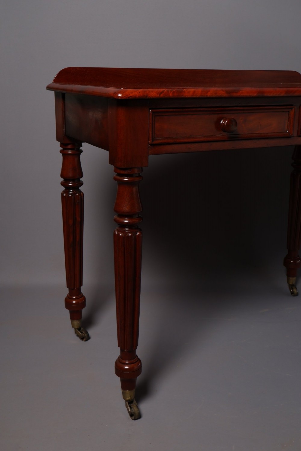 A Victorian mahogany writing table with a low gallery back, two frieze drawers with wooden knob handles, and turned reeded legs on brass castors. View IV
