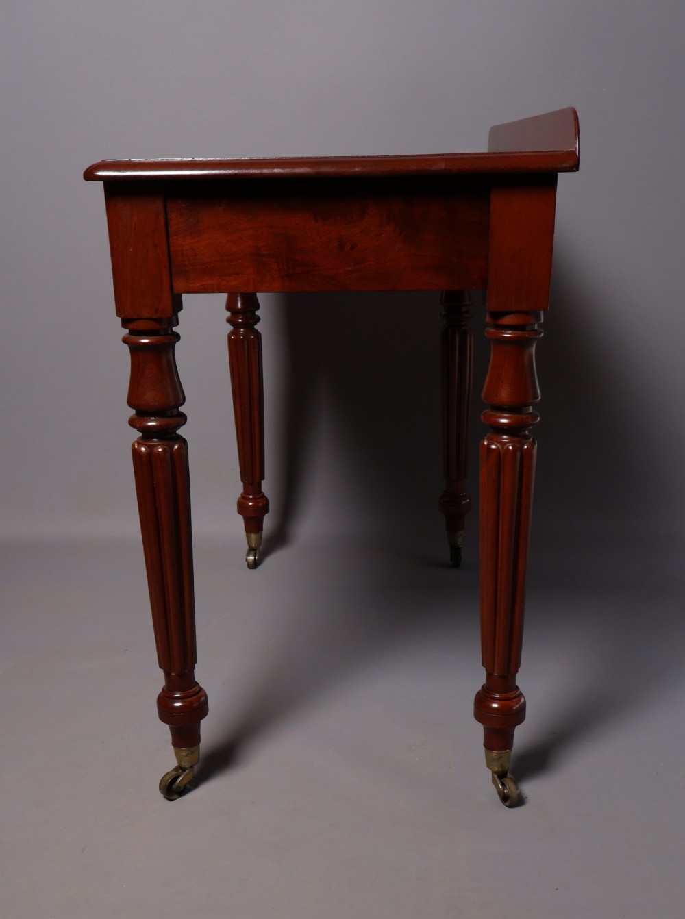 A Victorian mahogany writing table with a low gallery back, two frieze drawers with wooden knob handles, and turned reeded legs on brass castors. View VI