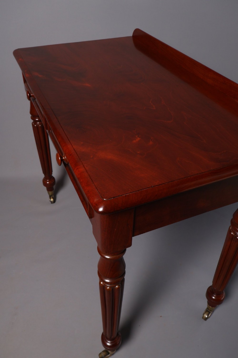 A Victorian mahogany writing table with a low gallery back, two frieze drawers with wooden knob handles, and turned reeded legs on brass castors. View VIII