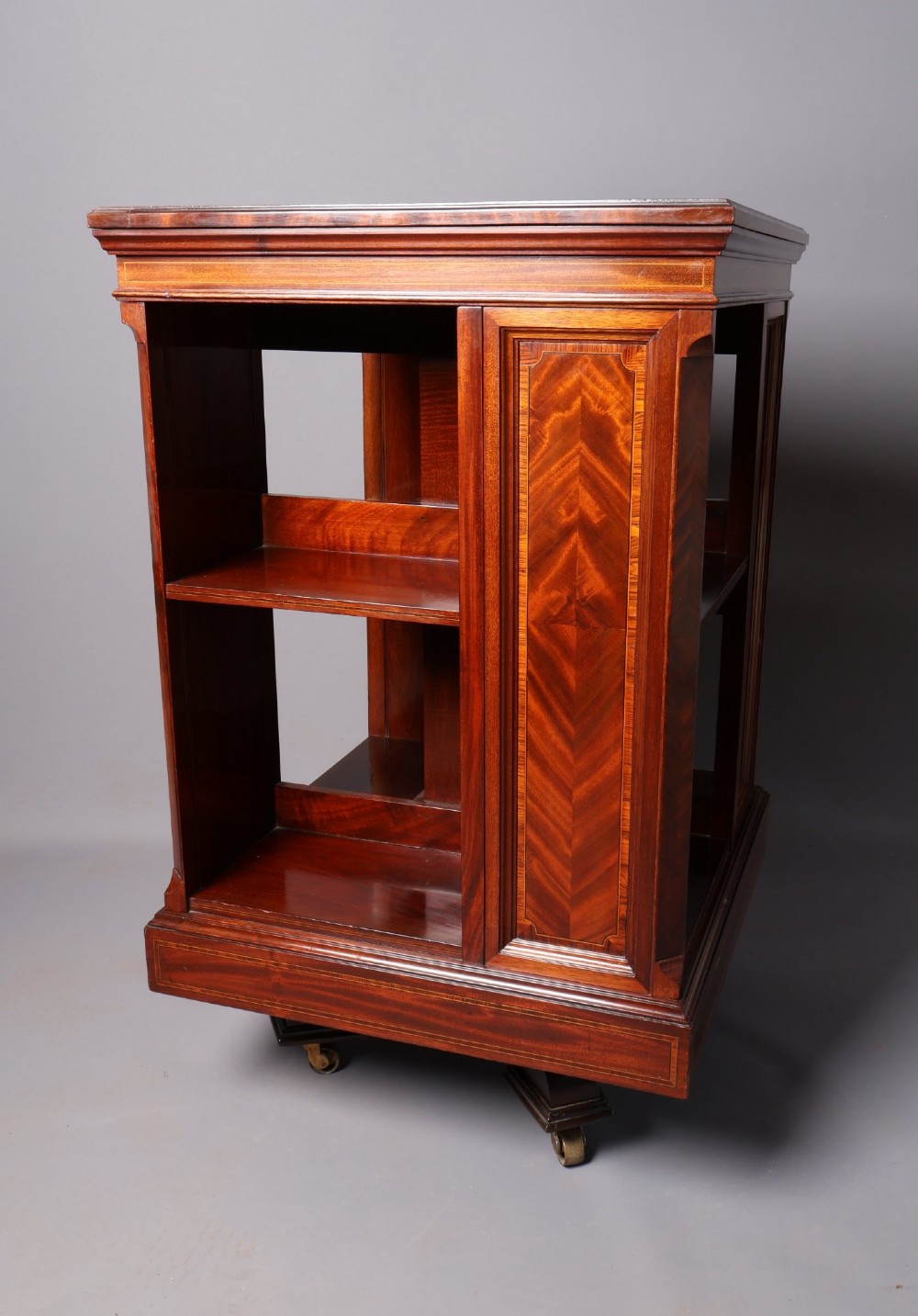 An Edwardian mahogany revolving bookcase featuring fine line inlay, flame-veneered vertical side panels, two shelving tiers, and a quadruped base with brass castors.