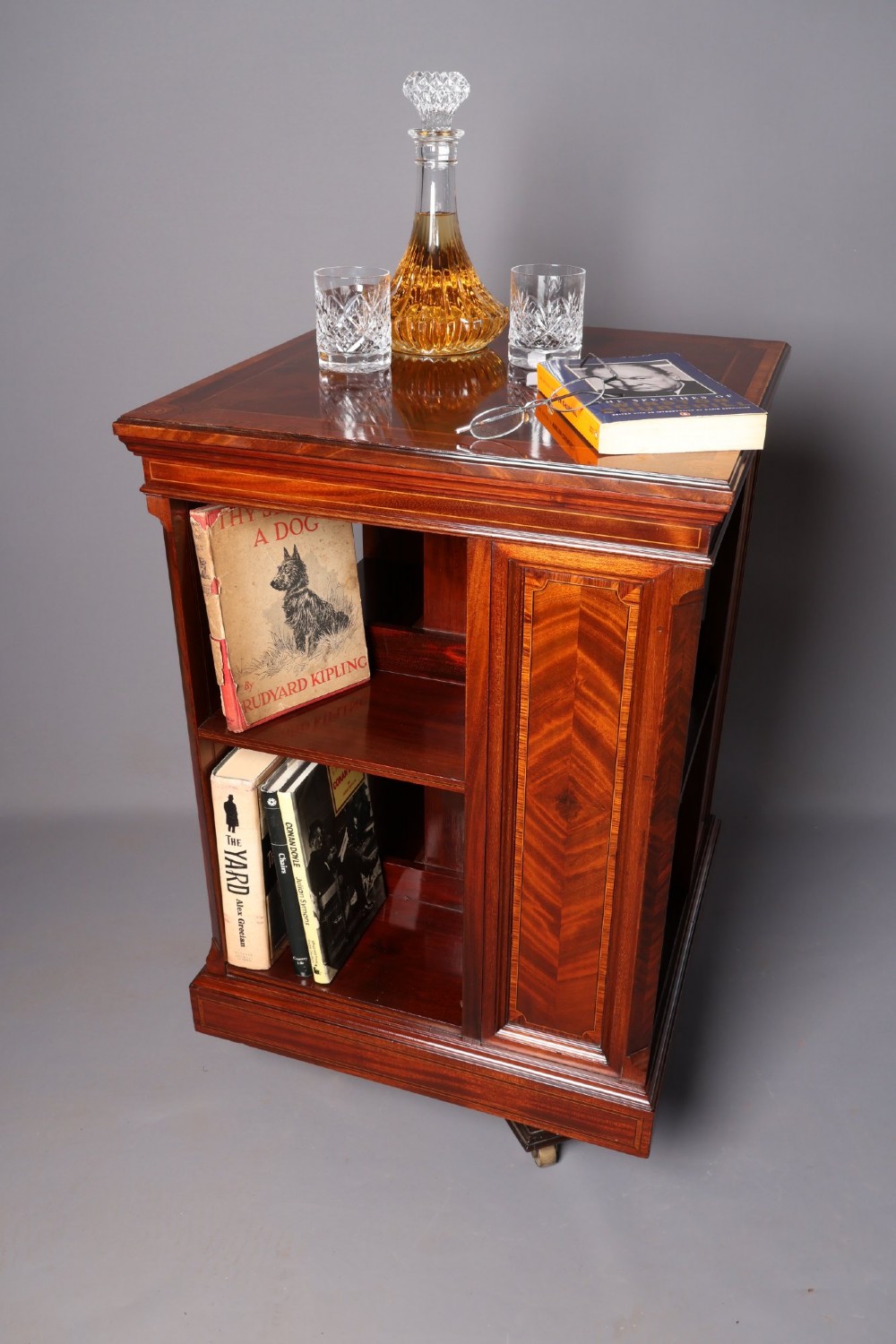 An Edwardian mahogany revolving bookcase featuring fine line inlay, flame-veneered vertical side panels, two shelving tiers, and a quadruped base with brass castors. View II
