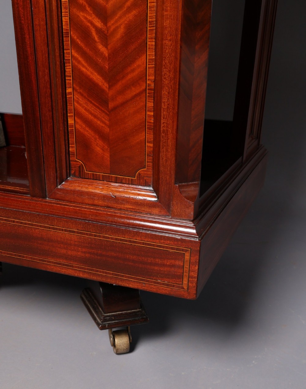 An Edwardian mahogany revolving bookcase featuring fine line inlay, flame-veneered vertical side panels, two shelving tiers, and a quadruped base with brass castors. View V