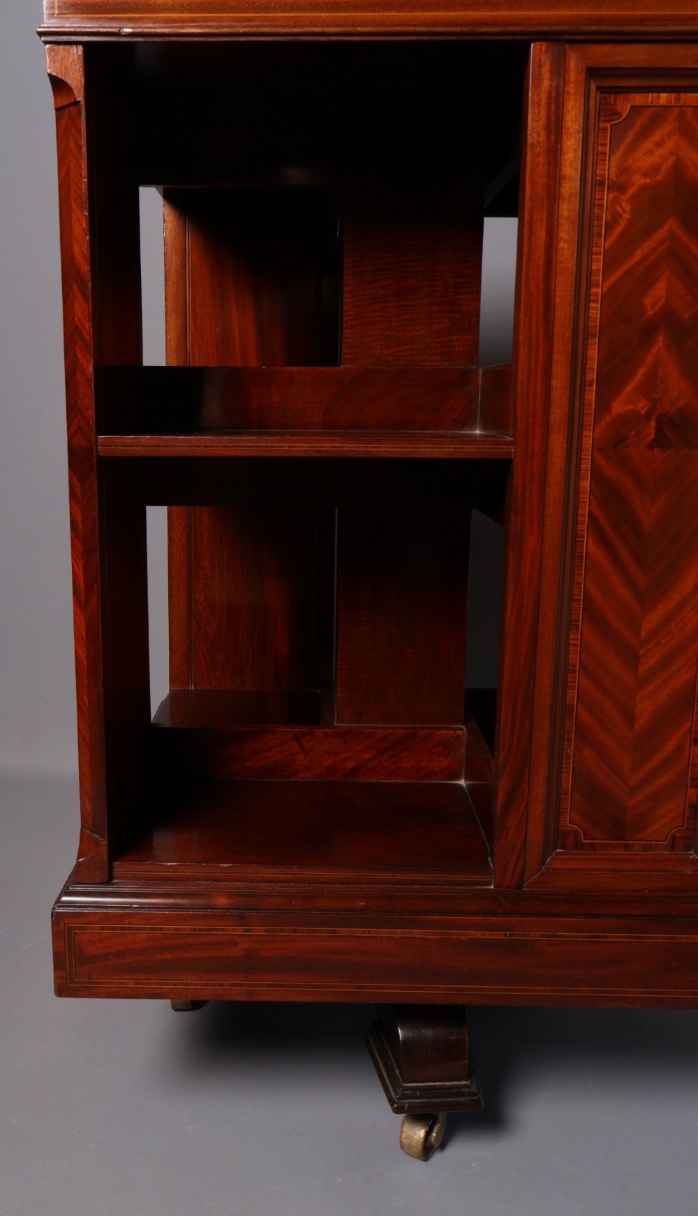 An Edwardian mahogany revolving bookcase featuring fine line inlay, flame-veneered vertical side panels, two shelving tiers, and a quadruped base with brass castors. View VI