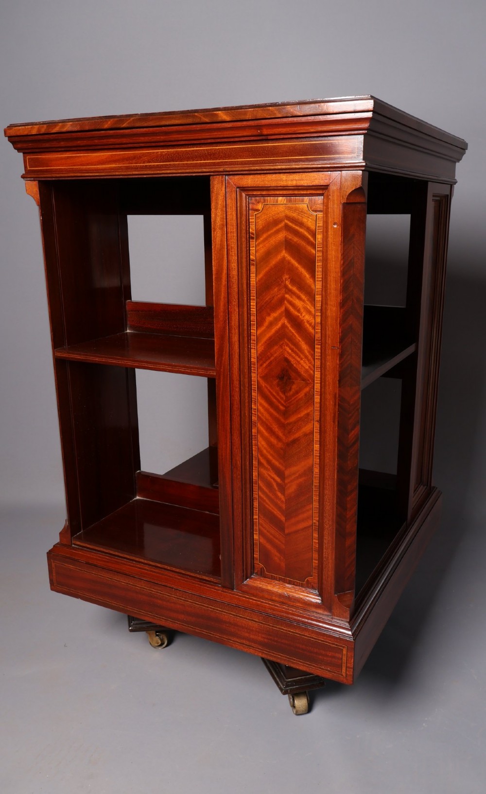 An Edwardian mahogany revolving bookcase featuring fine line inlay, flame-veneered vertical side panels, two shelving tiers, and a quadruped base with brass castors. View VII