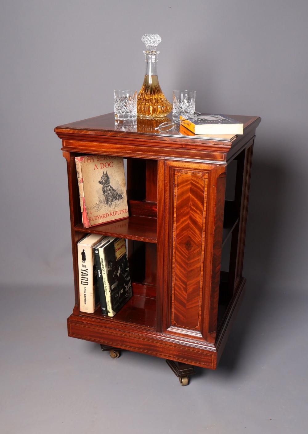 An Edwardian mahogany revolving bookcase featuring fine line inlay, flame-veneered vertical side panels, two shelving tiers, and a quadruped base with brass castors. View IX