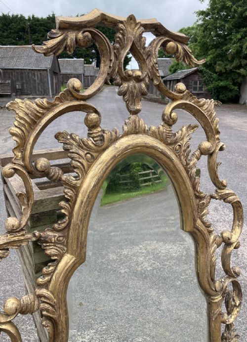 fig 1. A tall, narrow wall mirror framed in carved gold giltwood featuring elaborate acanthus leaf scrollwork and an ornate cresting.