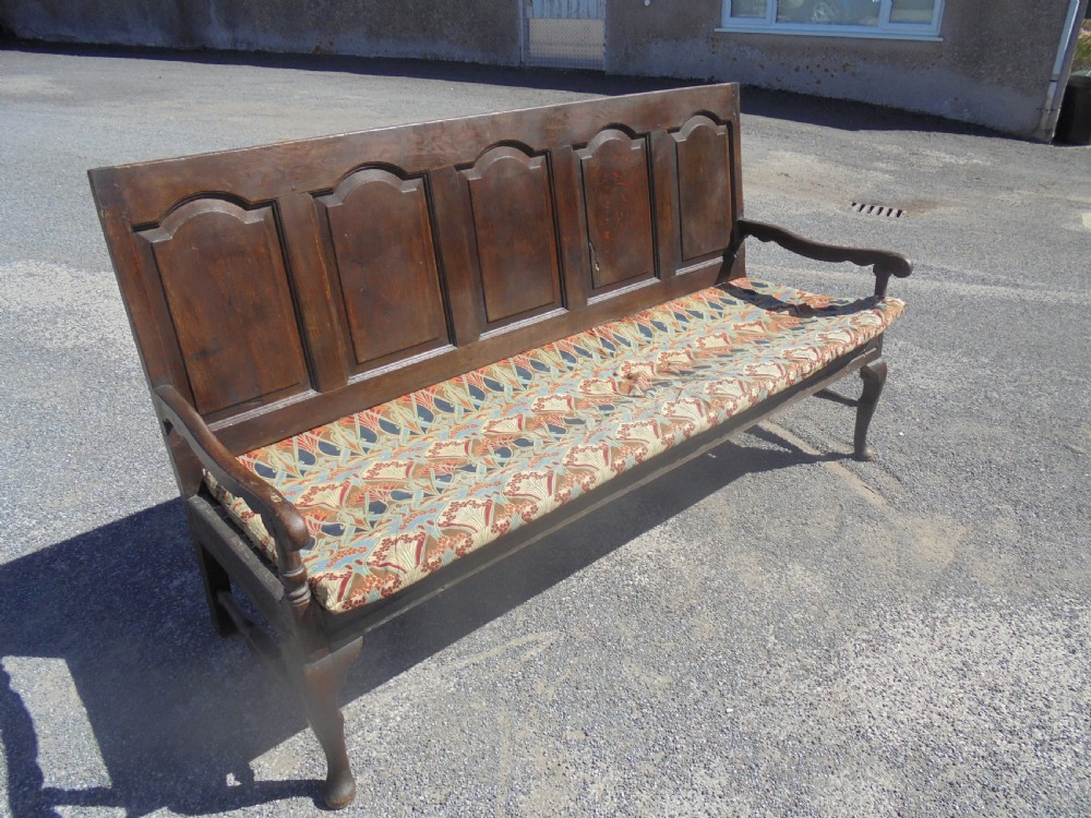 A dark oak wooden settle featuring five raised arched back panels, open curved armrests, cabriole front legs, and a patterned fabric seat cushion.
