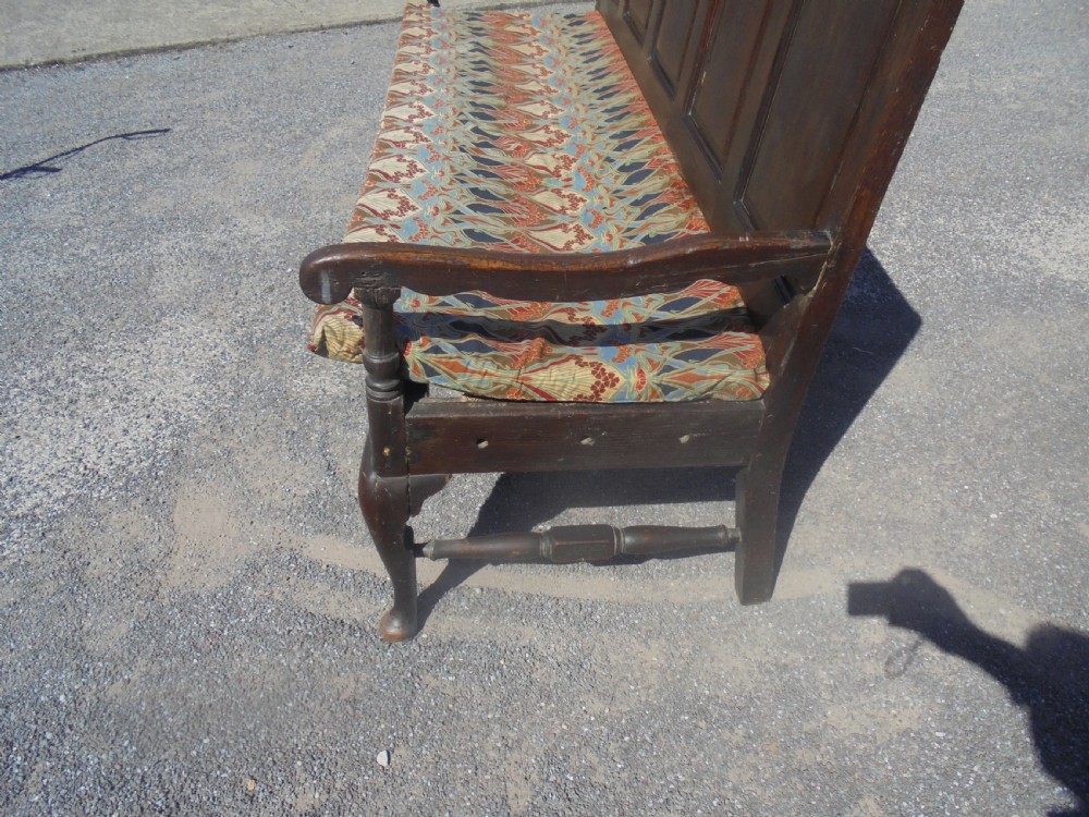 A dark oak wooden settle featuring five raised arched back panels, open curved armrests, cabriole front legs, and a patterned fabric seat cushion. View III