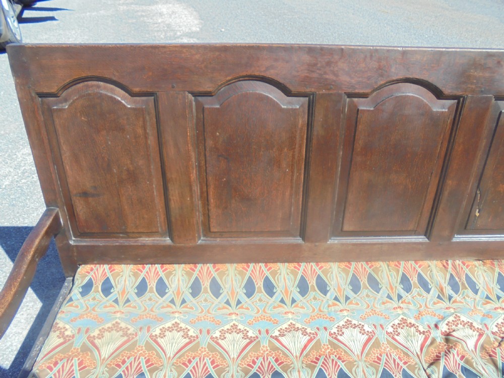 A dark oak wooden settle featuring five raised arched back panels, open curved armrests, cabriole front legs, and a patterned fabric seat cushion. View V