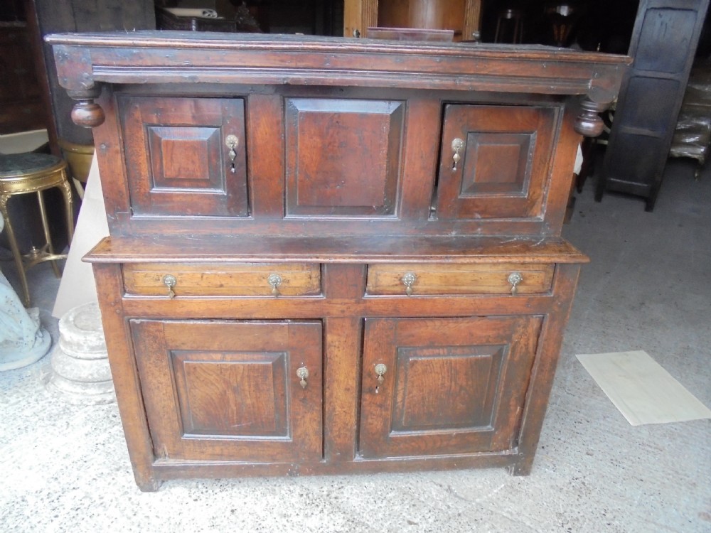 An early dark oak two-tier press cupboard featuring double geometric panelled doors, two drawers with brass teardrop handles, and turned drop finials.