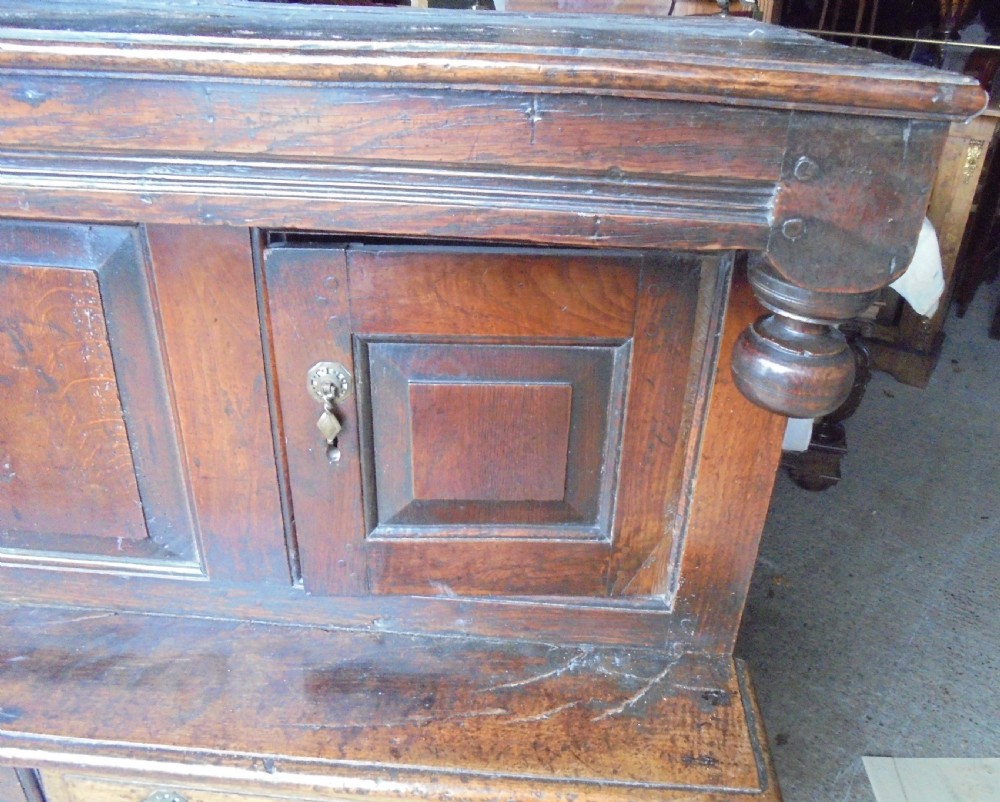 An early dark oak two-tier press cupboard featuring double geometric panelled doors, two drawers with brass teardrop handles, and turned drop finials. View II