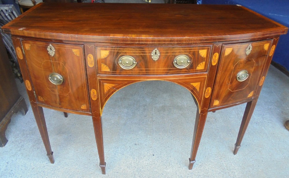 An 18th-century Sheraton style bowfront mahogany sideboard featuring satinwood inlays, brass drop handles, and square tapered legs.