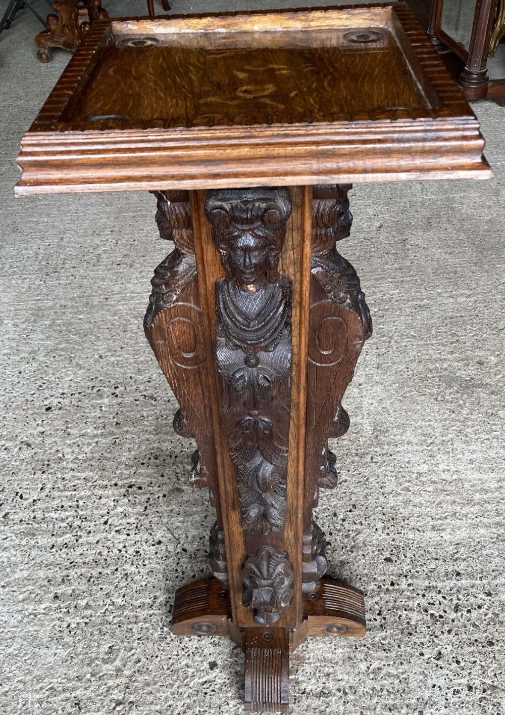 A carved dark oak torchere stand featuring ornate figural corner supports, a moulded square top, and a stepped base.