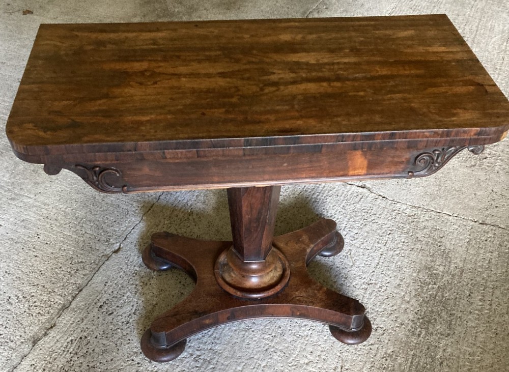 An early Victorian dark rosewood card table featuring a rectangular fold-over top, carved apron corner brackets, a central pedestal column, and a four-pointed platform base with bun feet.