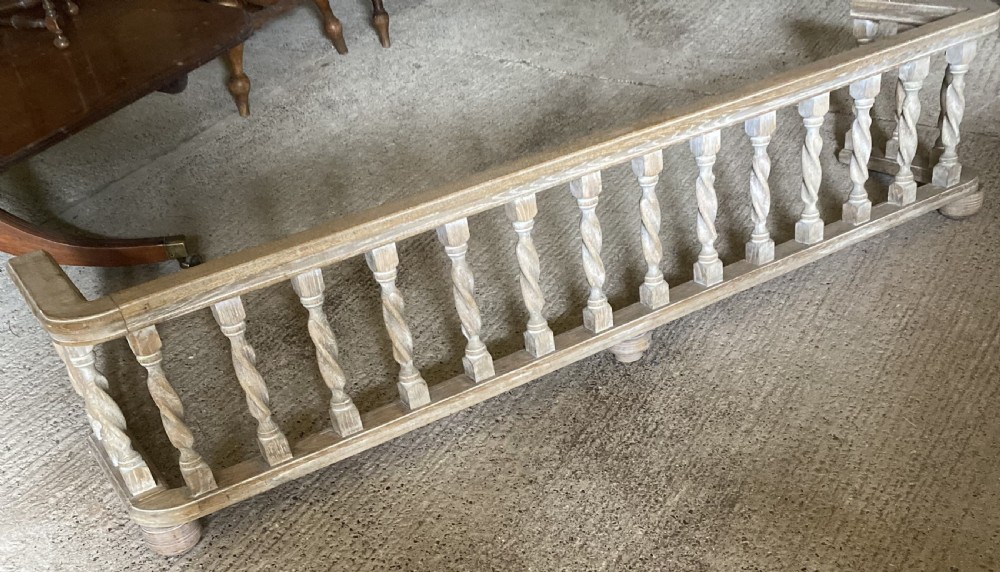 A wide limed oak fire fender featuring a row of twisted barley spindle balusters set between a top rail and bottom frame.