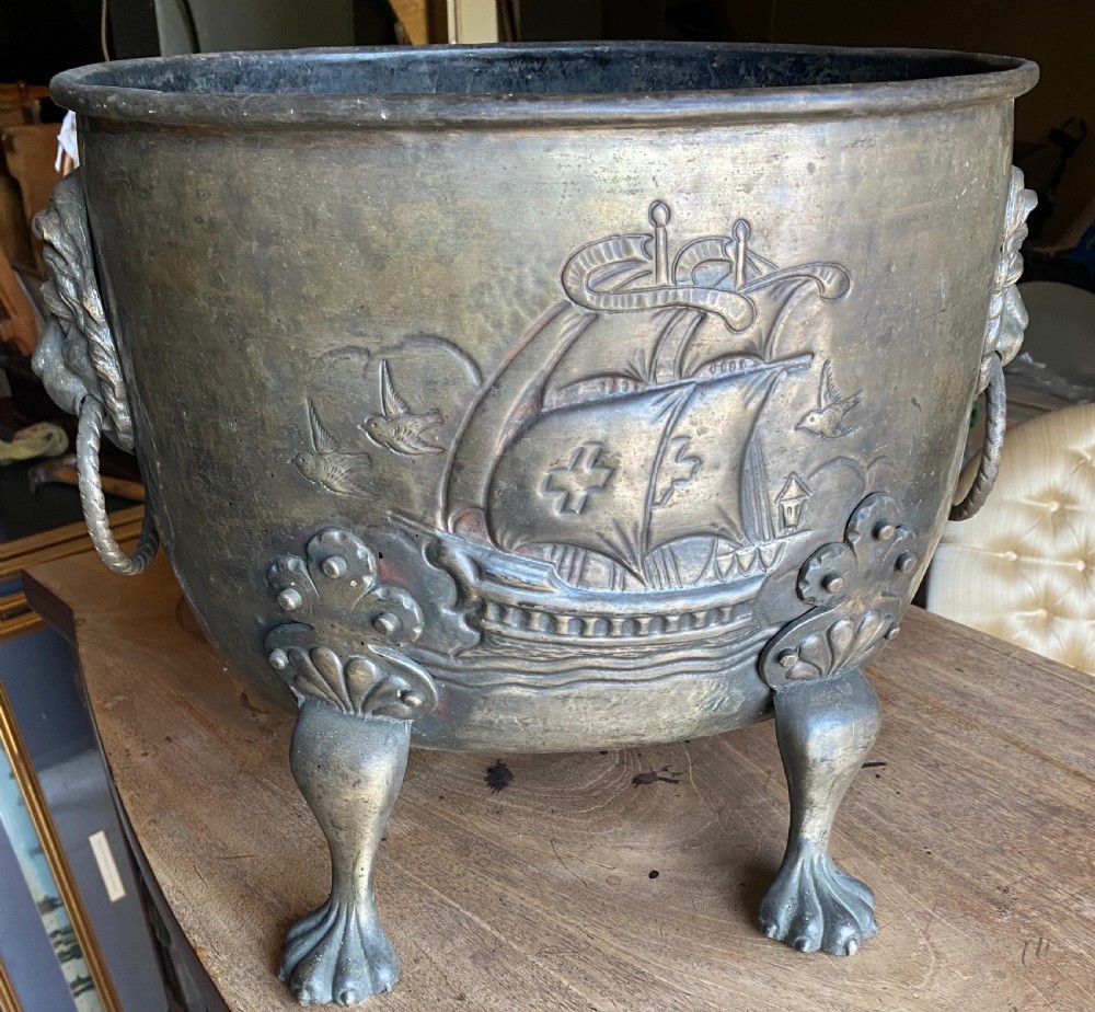 A deep bronzed brass log bucket decorated with a relief depiction of a sailing ship, supported by claw feet and featuring lion mask side handles.