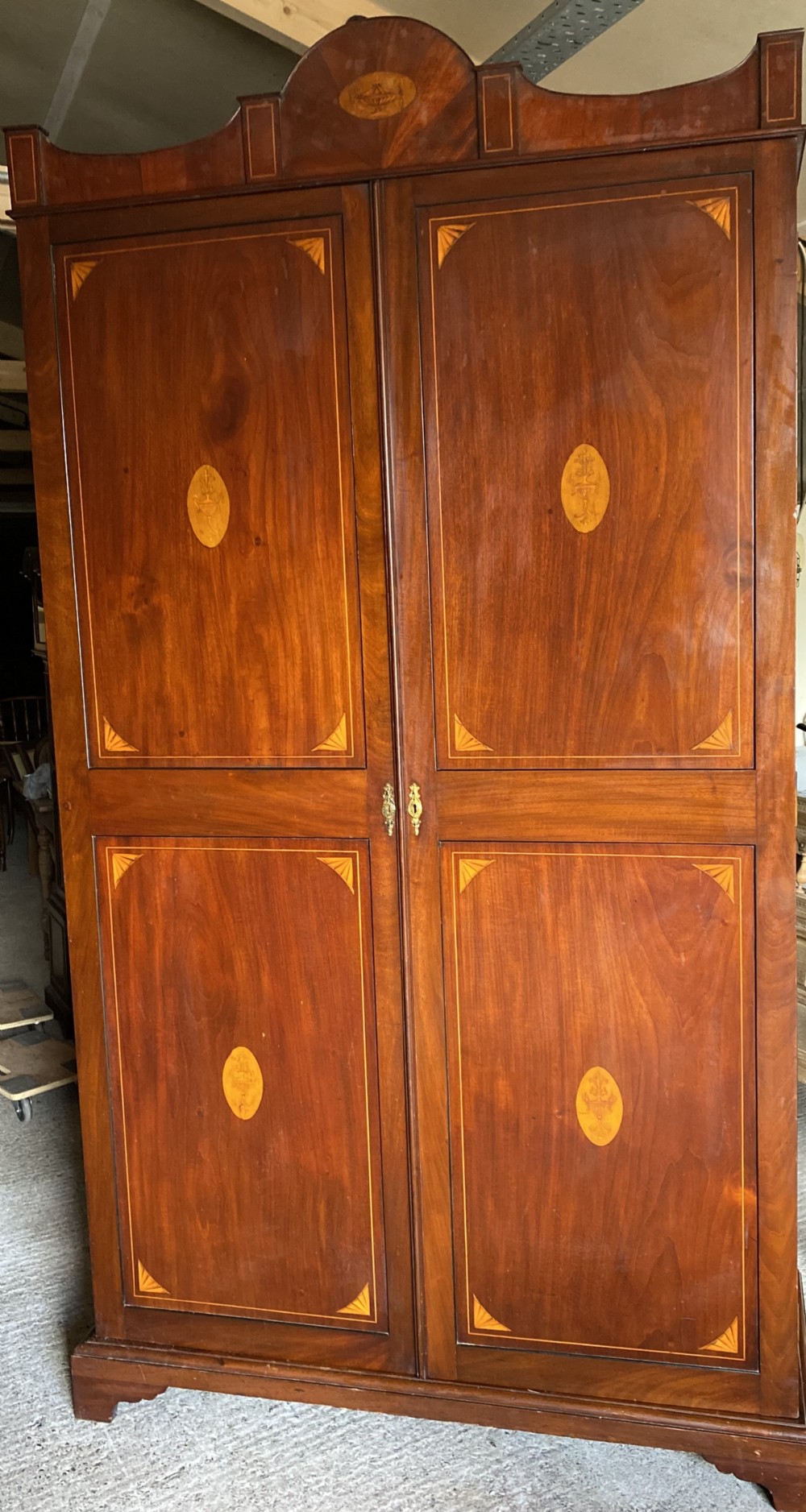A tall Sheraton style mahogany double wardrobe with twin panelled doors adorned with satinwood urn inlays and a shaped pediment.