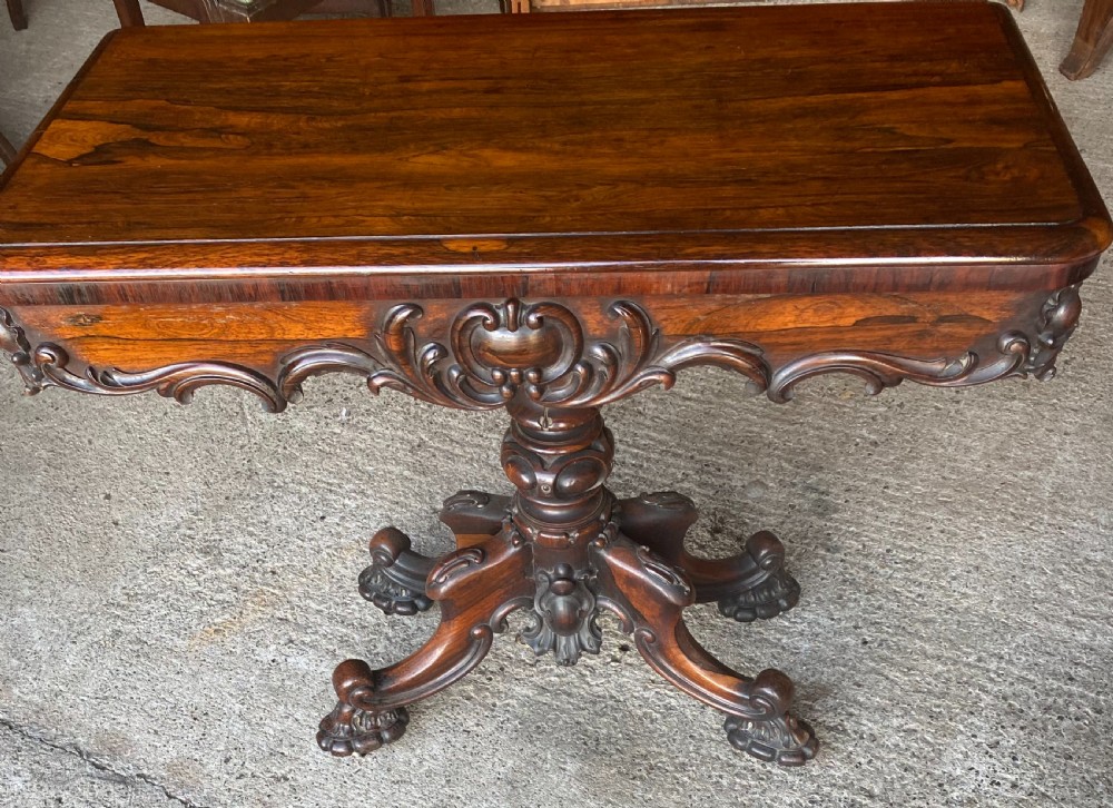 A Victorian rosewood fold-over card table featuring an intricately carved apron, turned central pedestal column, and four scrolled carved legs.