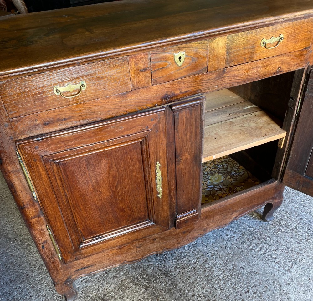An early 18th-century French cedarwood dresser base featuring two panelled cupboard doors, long exterior brass hinges, upper drawers, and a warm brown patinated timber finish. View II