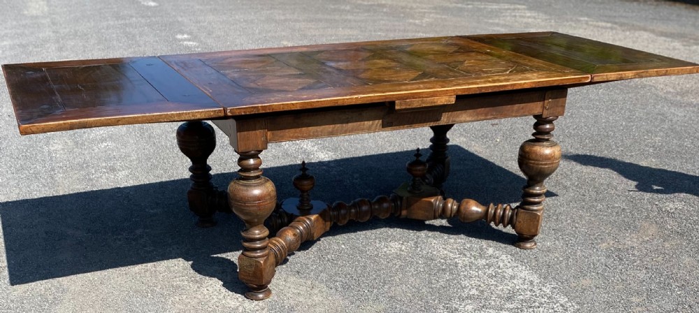 An 18th century Spanish fruitwood refectory dining table featuring a geometric parquet tabletop, draw-leaf extension panels, turned baluster legs, and a carved central stretcher.