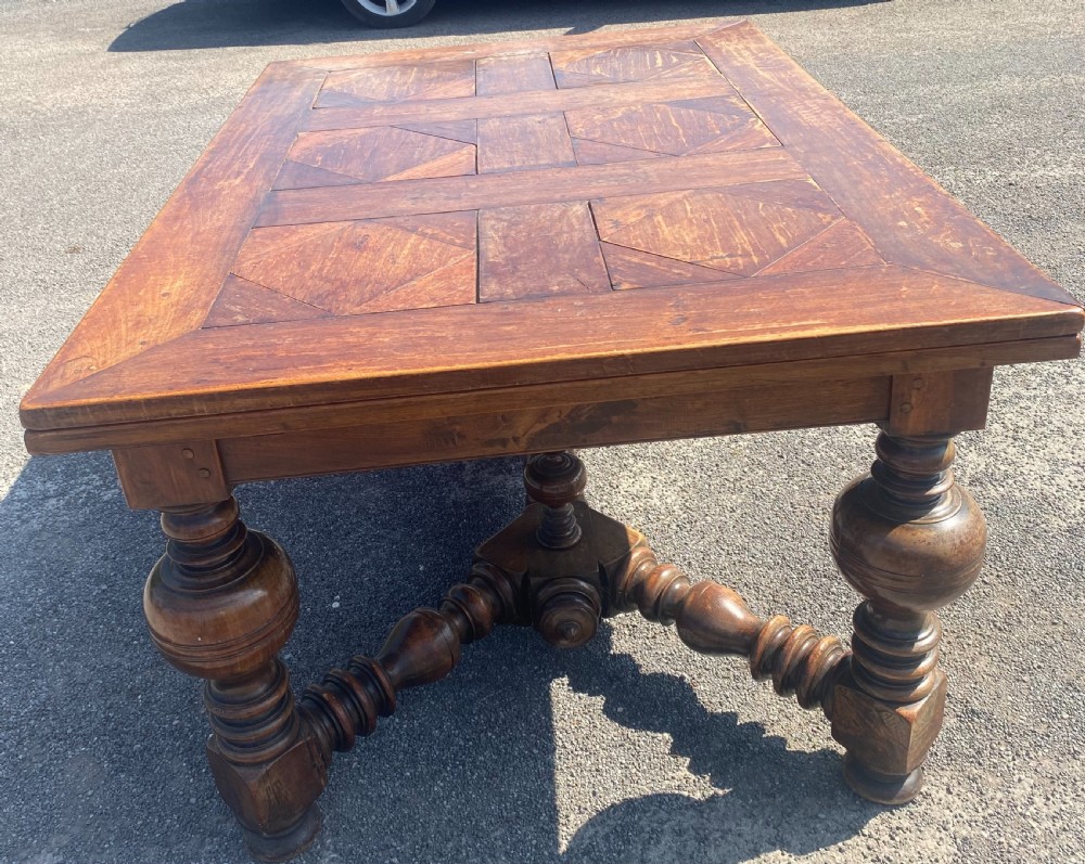 An 18th century Spanish fruitwood refectory dining table featuring a geometric parquet tabletop, draw-leaf extension panels, turned baluster legs, and a carved central stretcher. View V