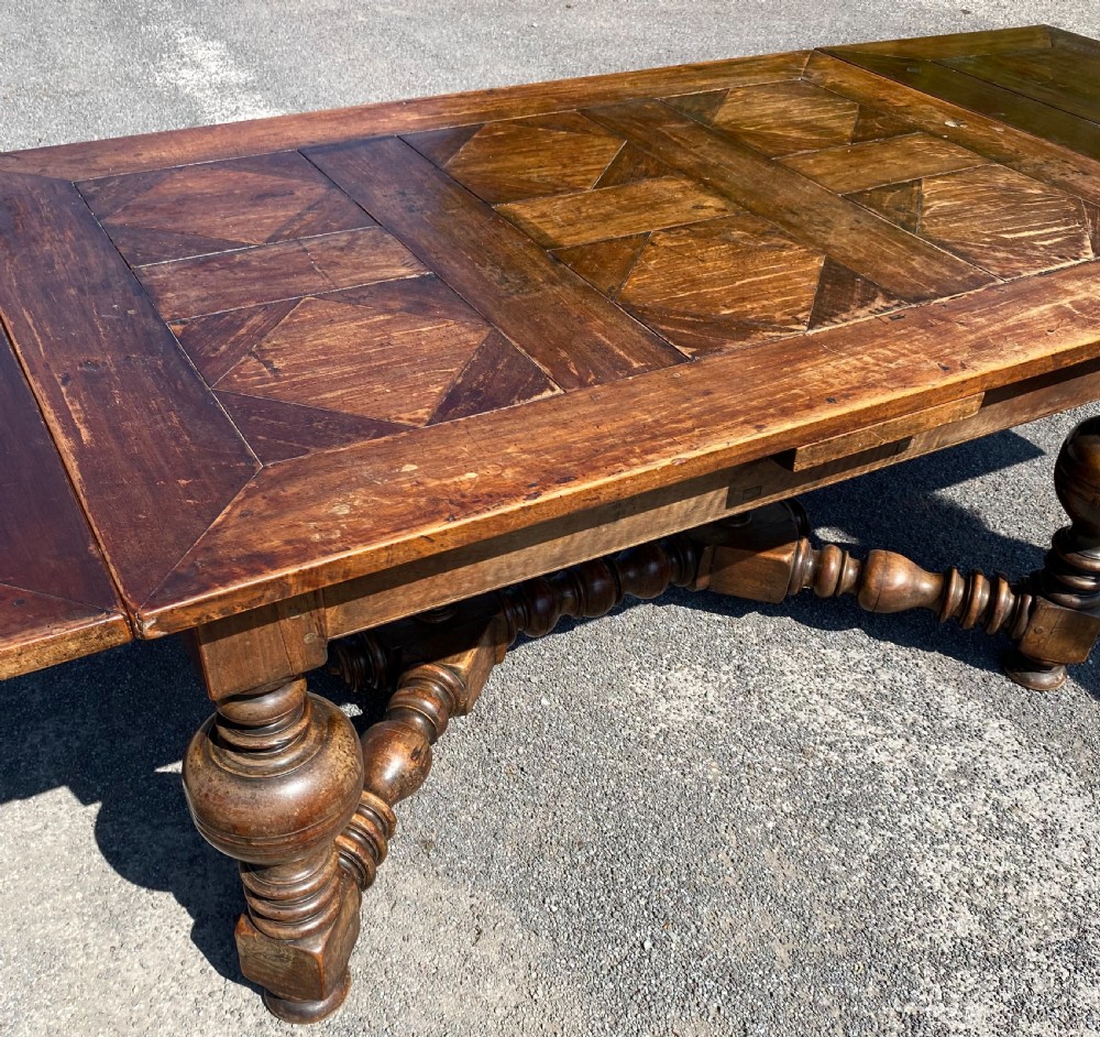 An 18th century Spanish fruitwood refectory dining table featuring a geometric parquet tabletop, draw-leaf extension panels, turned baluster legs, and a carved central stretcher. View X