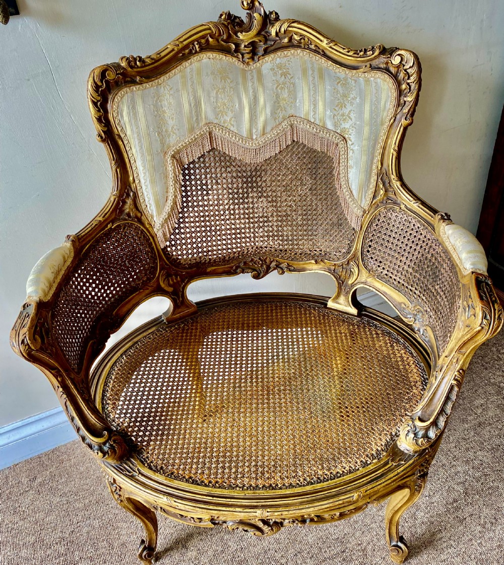 A 19th-century French giltwood bergere armchair featuring a carved ornate frame, woven cane seat and side panels, and a cream silk upholstered back section with tassel trim.