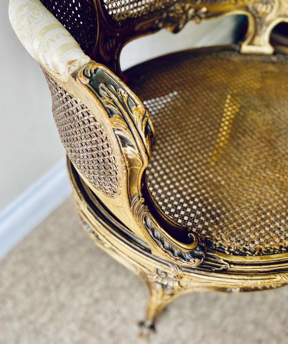 A 19th-century French giltwood bergere armchair featuring a carved ornate frame, woven cane seat and side panels, and a cream silk upholstered back section with tassel trim. View V