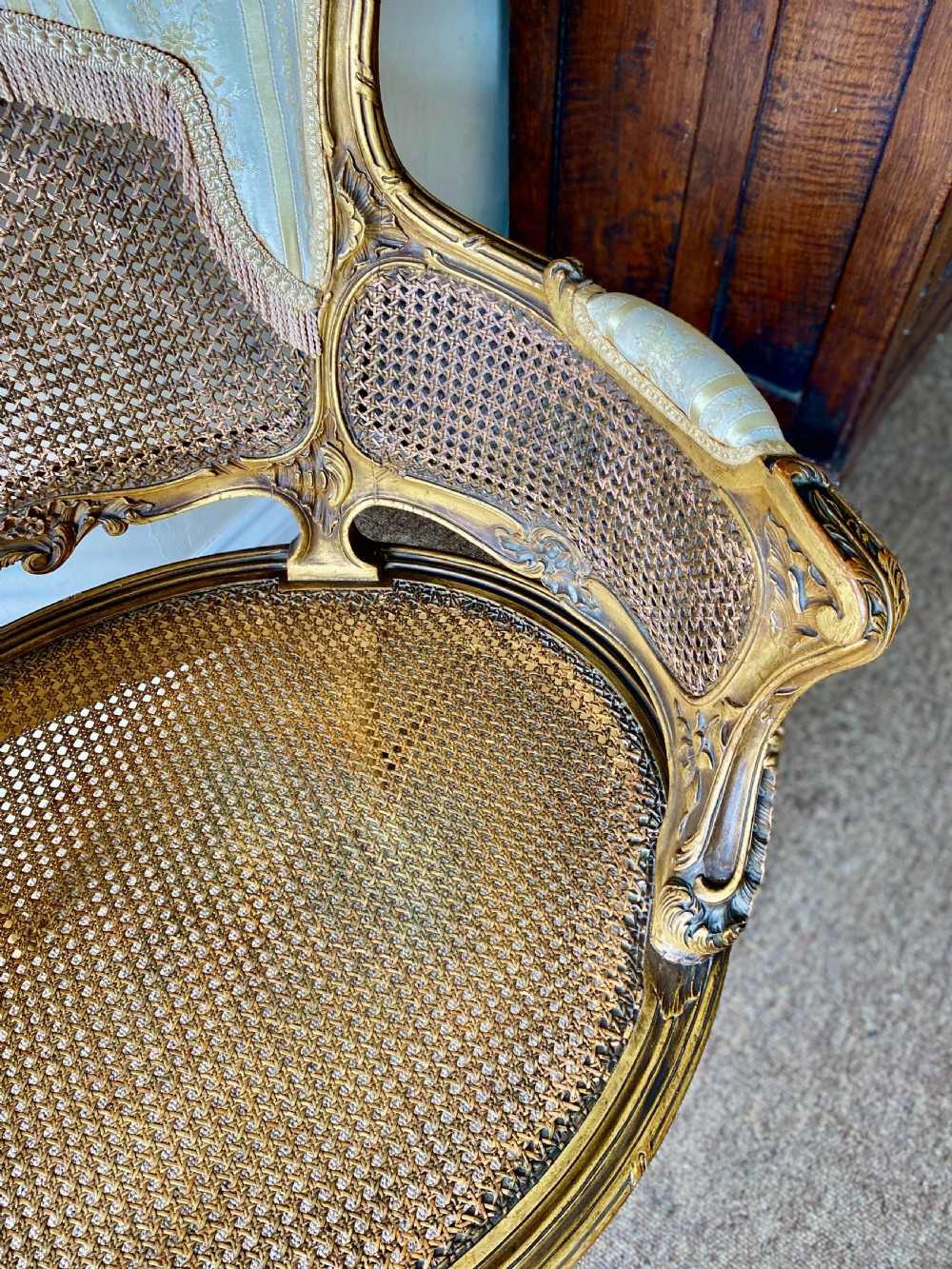 A 19th-century French giltwood bergere armchair featuring a carved ornate frame, woven cane seat and side panels, and a cream silk upholstered back section with tassel trim. View VI