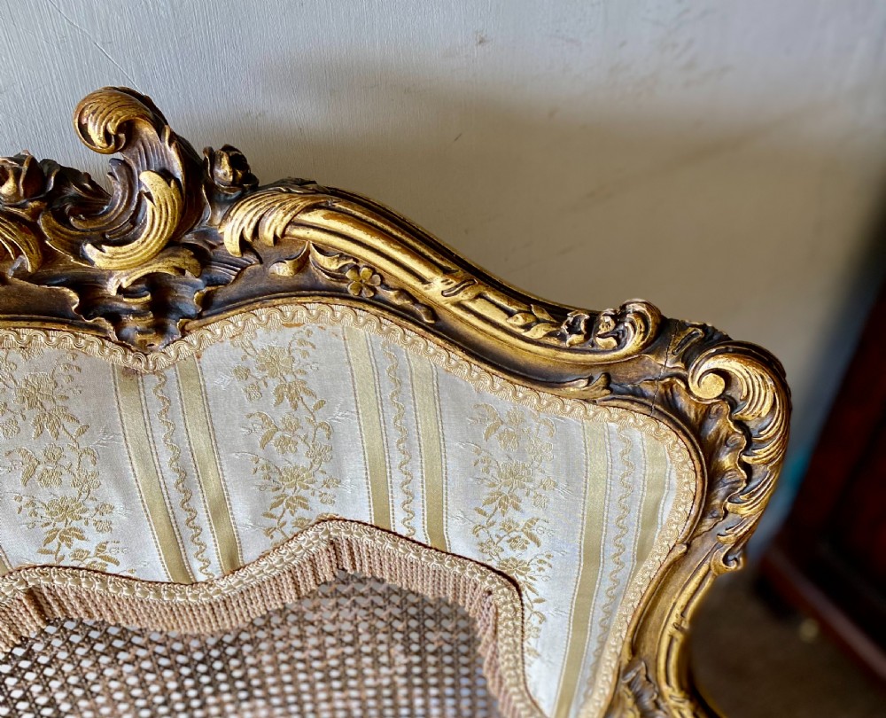 A 19th-century French giltwood bergere armchair featuring a carved ornate frame, woven cane seat and side panels, and a cream silk upholstered back section with tassel trim. View X