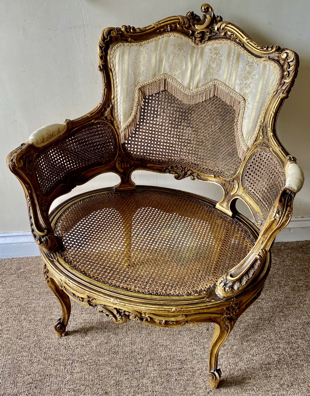 A 19th-century French giltwood bergere armchair featuring a carved ornate frame, woven cane seat and side panels, and a cream silk upholstered back section with tassel trim. View XI