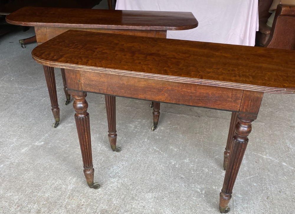 A pair of Regency mahogany console tables featuring rounded tops, reeded legs, and brass castors on a concrete surface.