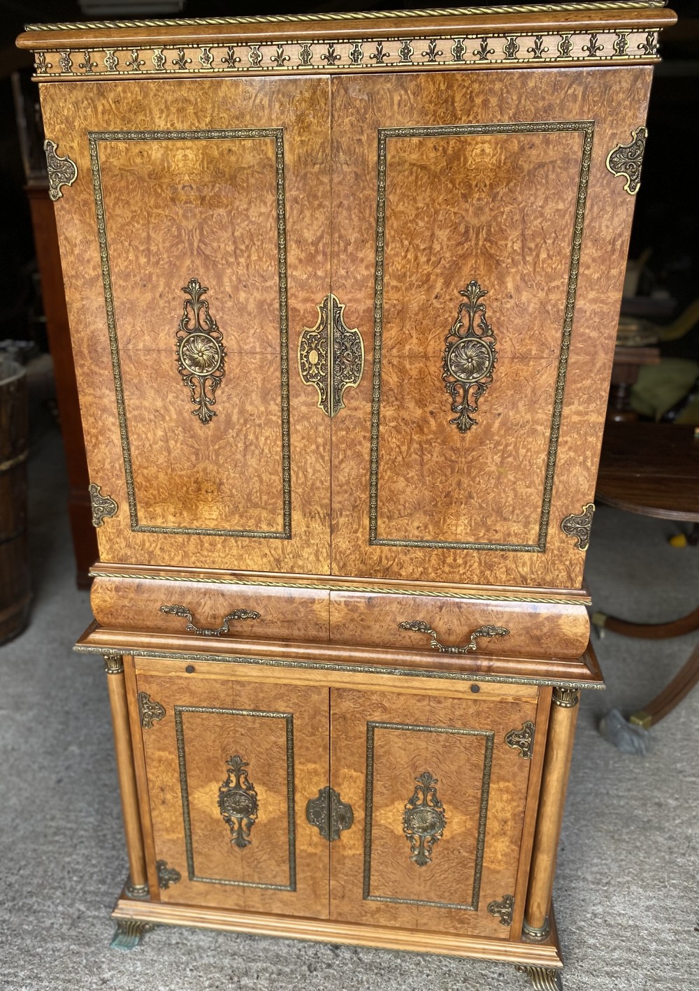 An ornate burr walnut cocktail cabinet featuring decorative brass mounts, central escutcheons, a middle drawer section, and twin lower cupboard doors.