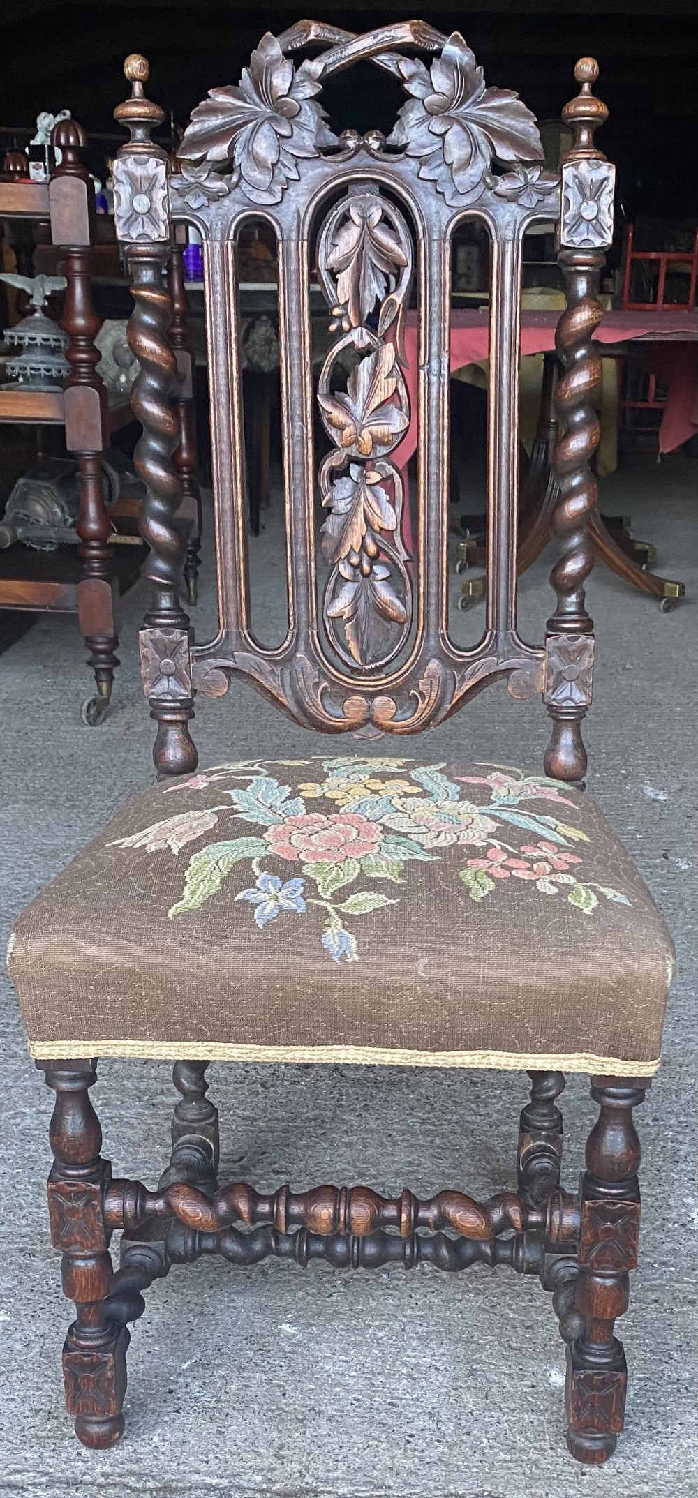 A dark carved oak dining chair featuring spiral turned legs, intricate leaf relief carvings on the backrest, and a beige upholstery seat with floral embroidery.