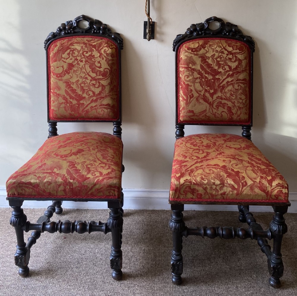 A pair of dark carved oak hall chairs featuring high backrests and seats upholstered in red and gold patterned damask fabric with turned legs.