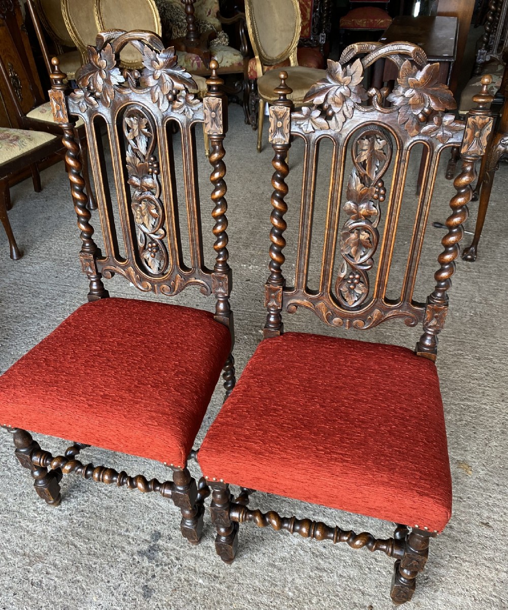 A pair of carved oak side chairs featuring high backrests with foliate carvings, barley twist uprights, and red textured upholstered seats.