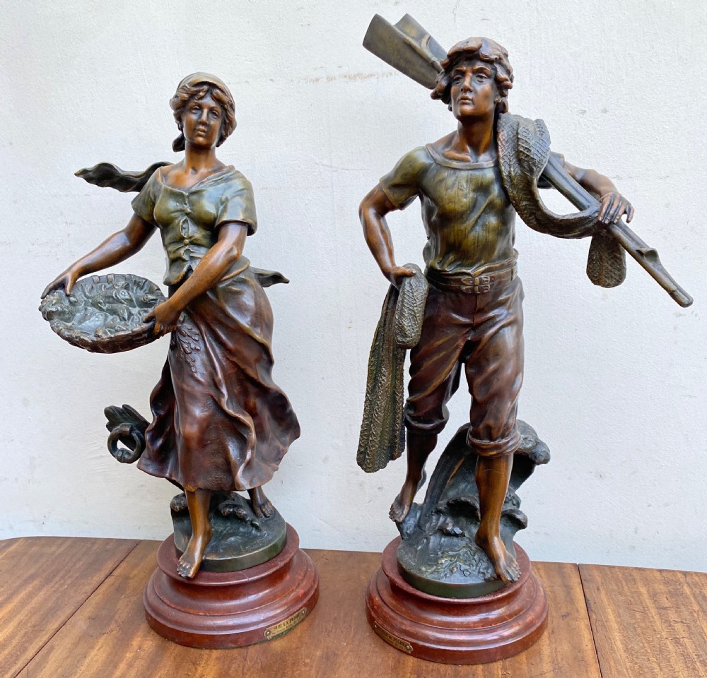 A pair of patinated spelter figures depicting a fisherwoman holding a basket and a fisherman carrying nets, mounted on circular turned wood bases with brass title plates.