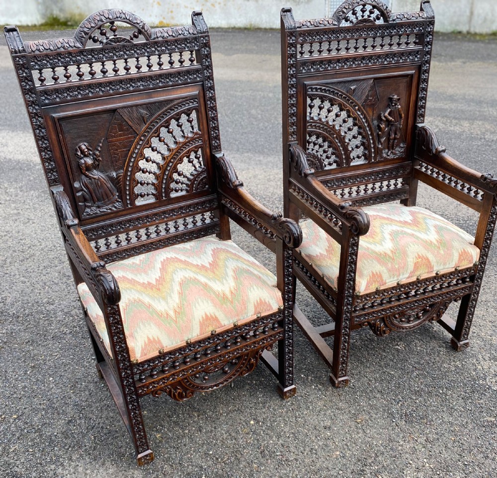 A pair of high-backed dark carved oak armchairs featuring intricate relief panels depicting folk figures, spindle galleries, and patterned fabric seats.