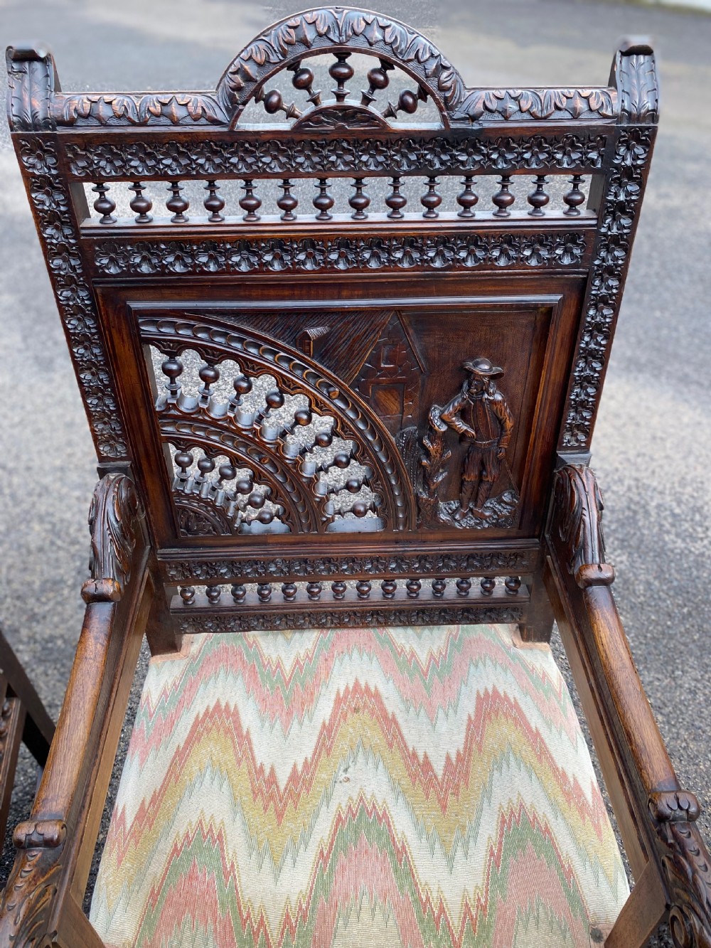 A pair of high-backed dark carved oak armchairs featuring intricate relief panels depicting folk figures, spindle galleries, and patterned fabric seats. View II