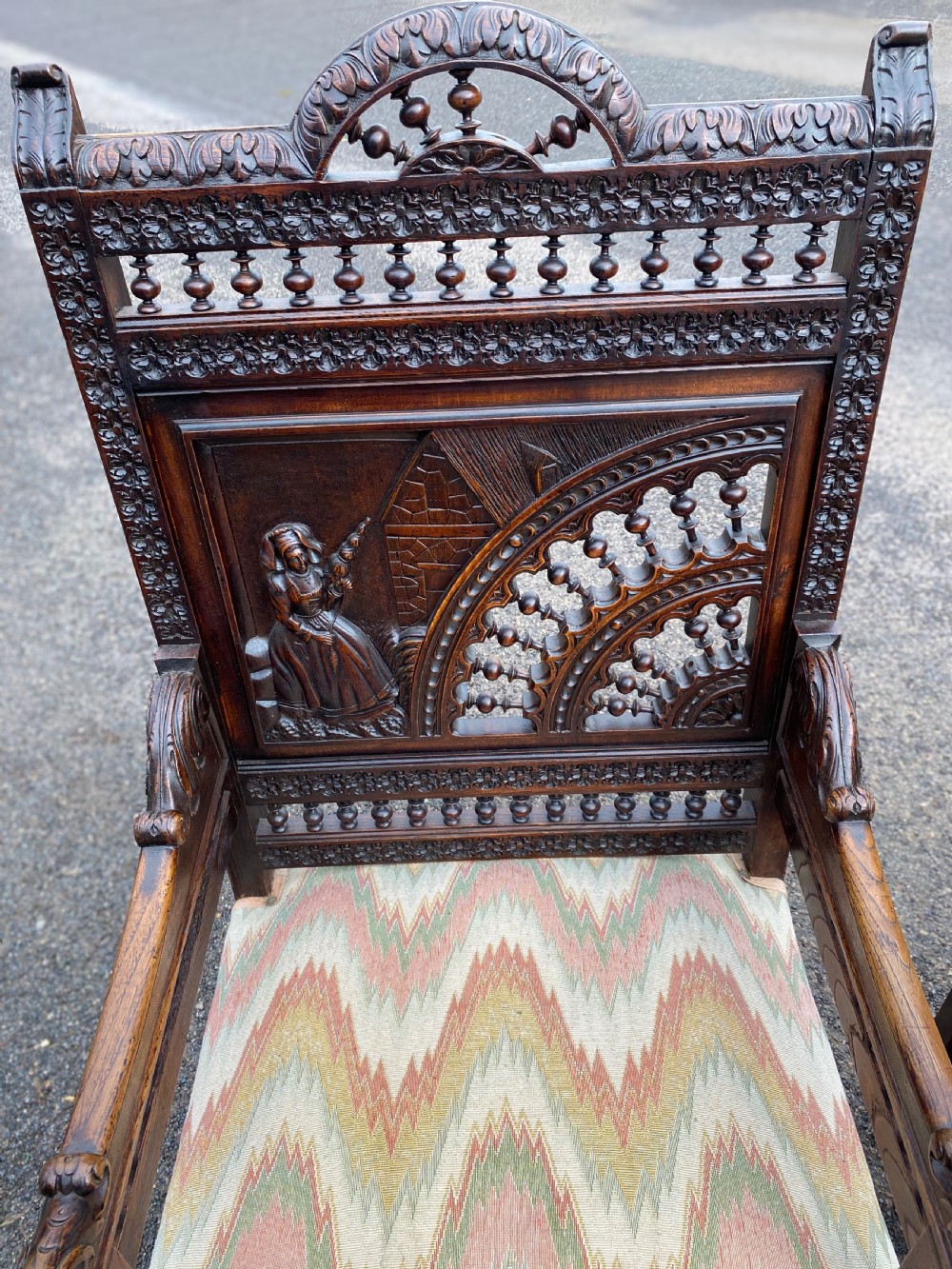 A pair of high-backed dark carved oak armchairs featuring intricate relief panels depicting folk figures, spindle galleries, and patterned fabric seats. View III