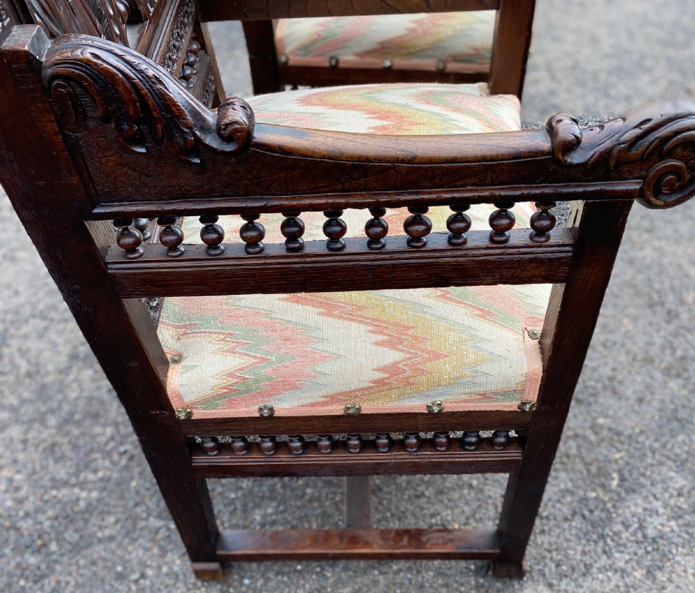 A pair of high-backed dark carved oak armchairs featuring intricate relief panels depicting folk figures, spindle galleries, and patterned fabric seats. View IV