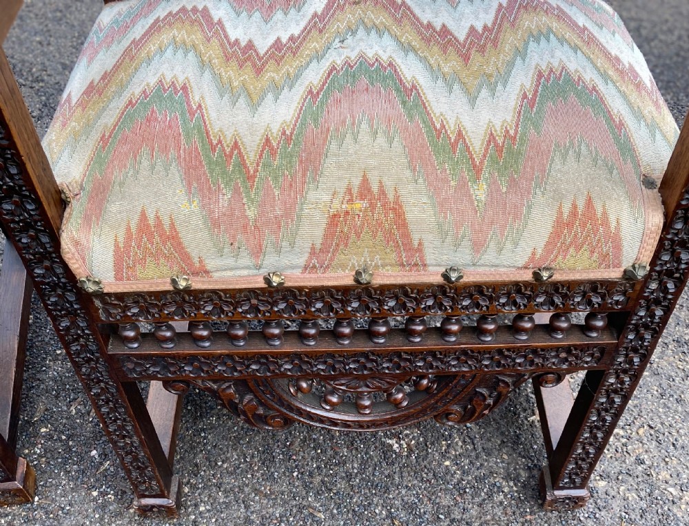 A pair of high-backed dark carved oak armchairs featuring intricate relief panels depicting folk figures, spindle galleries, and patterned fabric seats. View V