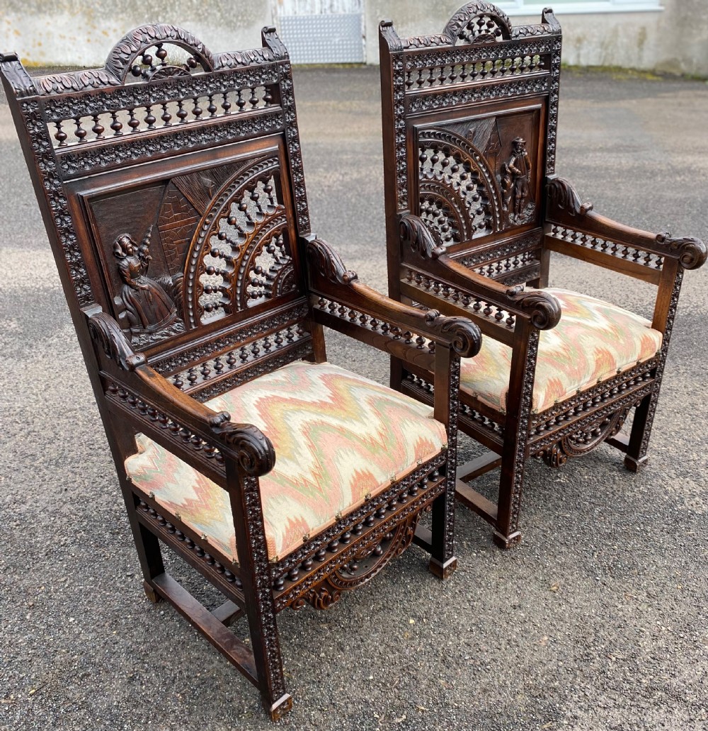 A pair of high-backed dark carved oak armchairs featuring intricate relief panels depicting folk figures, spindle galleries, and patterned fabric seats. View VI
