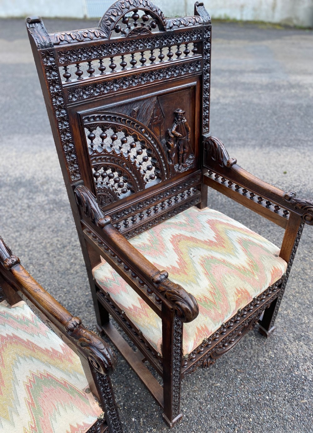 A pair of high-backed dark carved oak armchairs featuring intricate relief panels depicting folk figures, spindle galleries, and patterned fabric seats. View VII