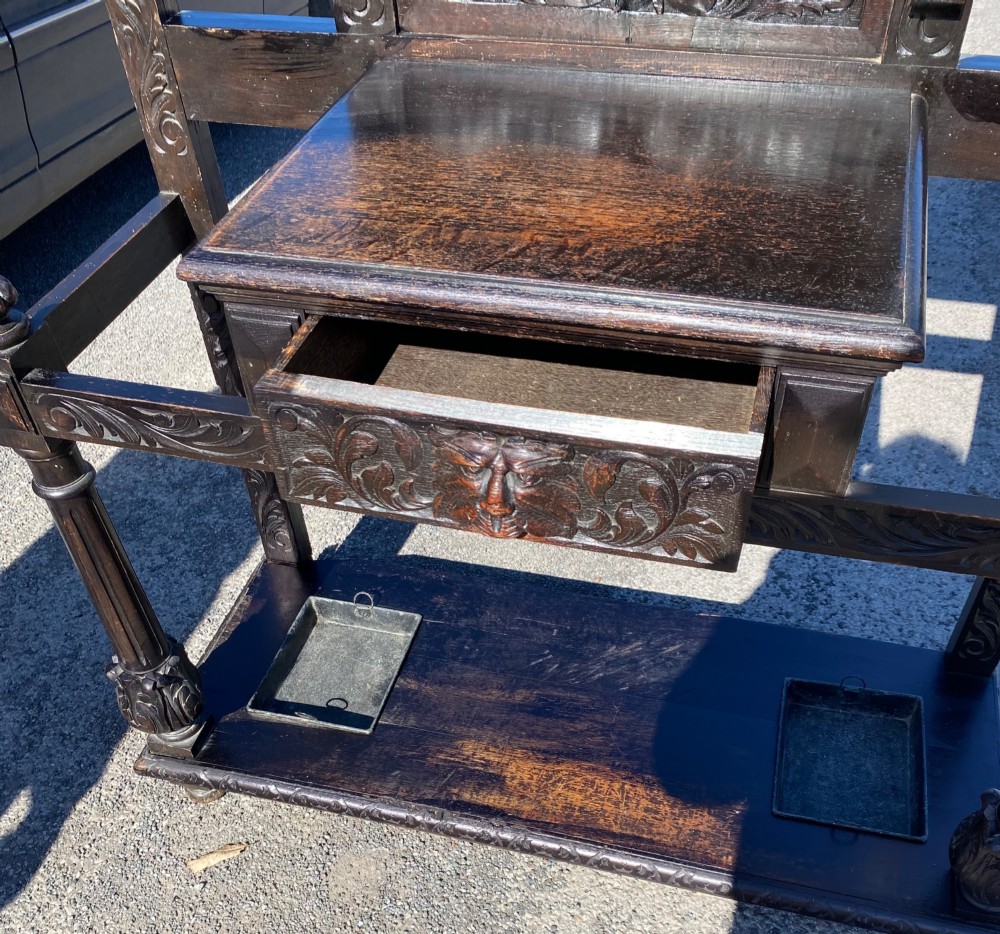A dark carved oak Victorian hall stand featuring a central rectangular mirror, lion mask carvings, turned coat pegs, umbrella holders, and metal drip trays. View V